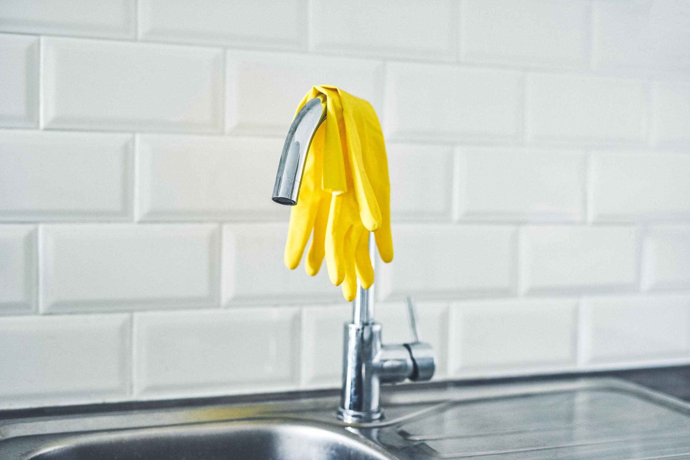 Clean Kitchen Sink with Yellow Rubber Gloves Hanging Off Sink Water Tap Against a White Tile Backsplash