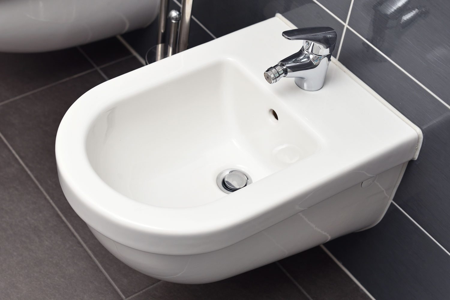 Close up of White Bidet Screwed To A Gray Tiled Wall In The Bathroom