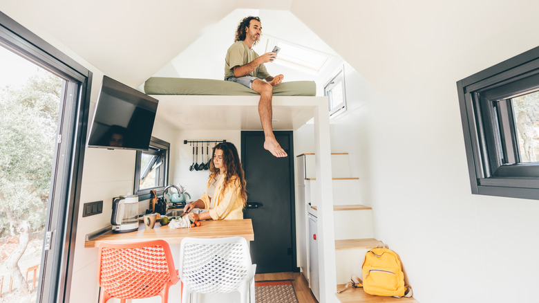 Une jeune femme travaille dans une cuisine tandis qu'un homme est assis au-dessus d'elle dans le grenier d'une petite maison