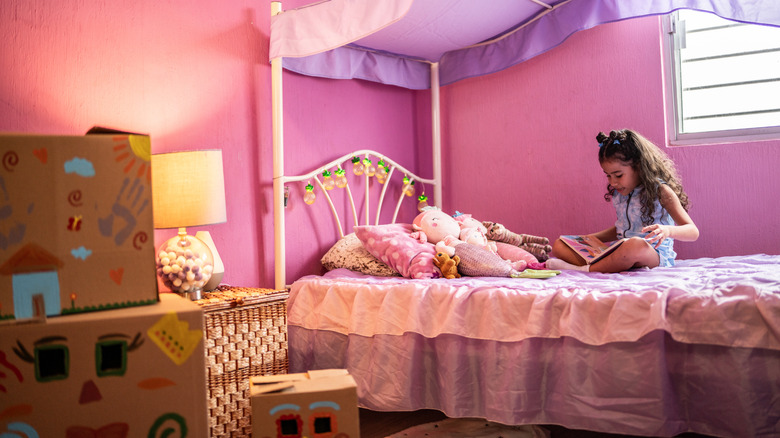 Une petite fille assise sur un lit de princesse rose avec des jouets à fourrure et lisant un livre.