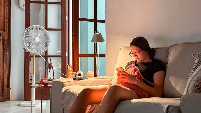 Un ventilateur stratégiquement placé montre une personne assise sur un canapé dans une pièce sombre