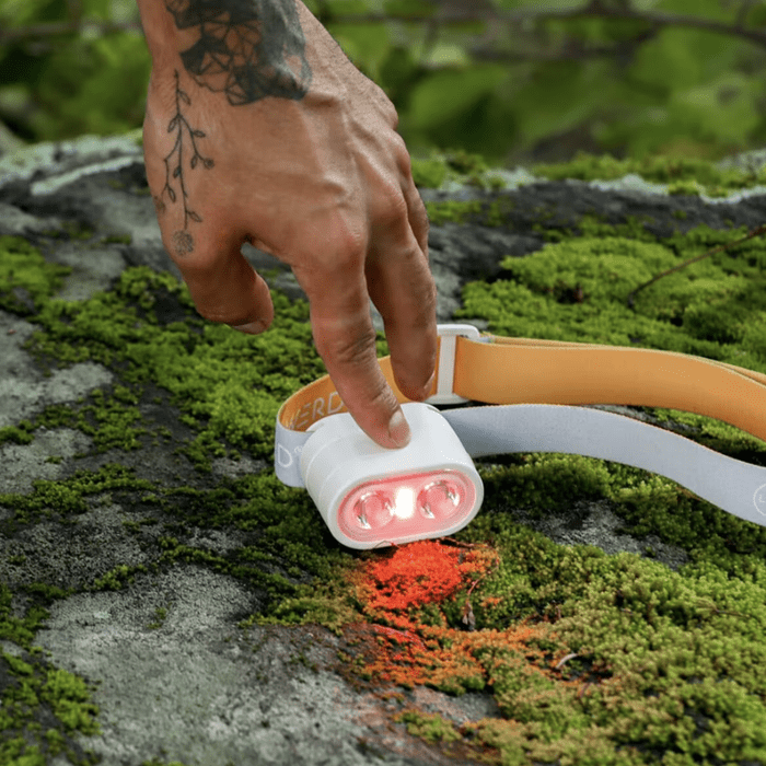 Une main touche un phare sur un rocher moussu, activant ses lumières dans un cadre extérieur naturel. Les couleurs vives contrastent avec la verdure.