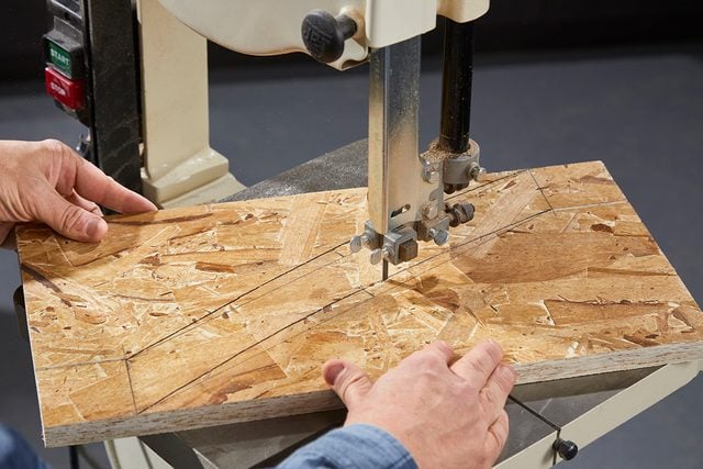 Une personne coupe un morceau de panneau à copeaux orientés (OSB) avec une scie à ruban, guidant soigneusement le bois le long des lignes marquées sous la lame de scie.
