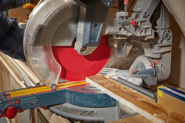 Gros plan d'une scie à onglets circulaire avec une lame rouge coupant un morceau de bois, avec de la sciure visible et des guides de mesure sur la surface de travail.