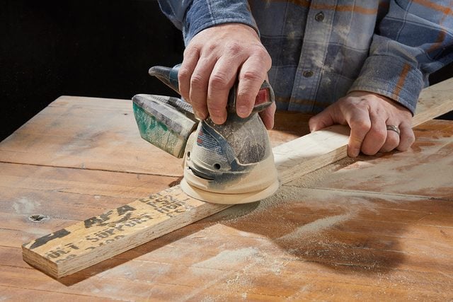 Une personne utilise une ponceuse électrique pour lisser une planche de bois sur un établi, vêtue d'une chemise à carreaux. De la poussière de bois est visible autour de la zone de ponçage.