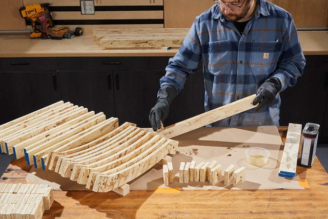 Une personne portant des gants et une chemise à carreaux dispose des pièces de bois courbées sur un établi, se préparant au travail du bois dans un atelier.