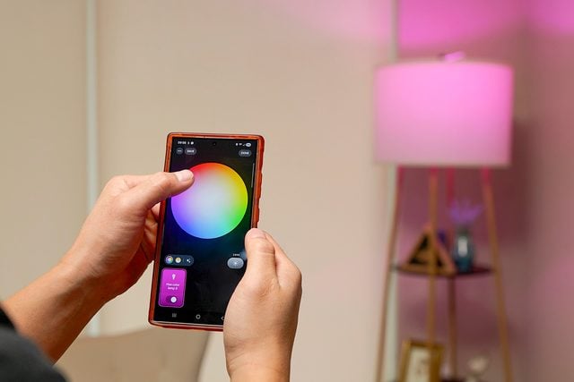 Une personne utilise une application pour smartphone pour régler la couleur d'une ampoule intelligente, changeant ainsi la lumière d'une lampe en rose dans un salon moderne.