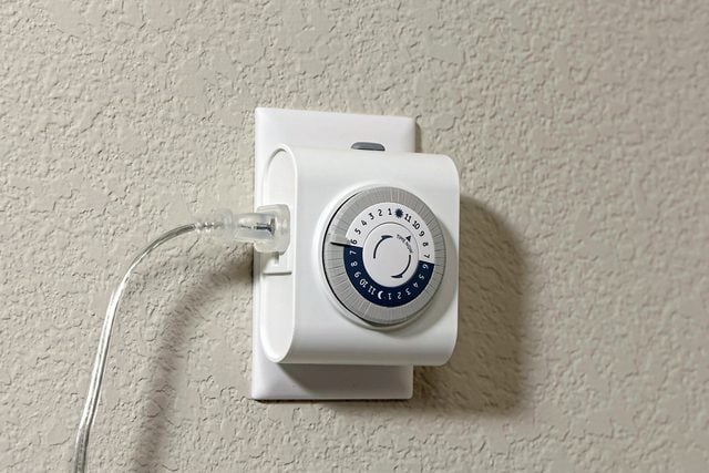 Une minuterie électrique blanche branchée sur une prise murale, avec un cordon transparent attaché. La minuterie a un cadran circulaire avec des chiffres noirs et de petits interrupteurs mécaniques sur le pourtour.