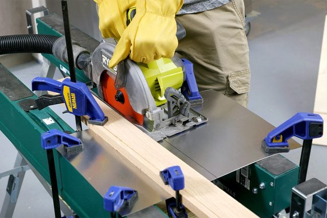 Une personne portant des gants jaunes utilise une scie circulaire pour couper un morceau de bois soutenu par des pinces sur un établi. Diverses pinces maintiennent le bois en place et la personne se tient au-dessus du banc, guidant soigneusement la scie le long du bois.