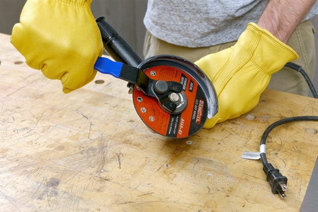 un gros plan d'une personne préparant une meuleuse d'angle à utiliser ; L'individu porte des gants de protection jaunes et semble ajuster ou fixer le disque de coupe sur la meuleuse d'angle ; La meuleuse d'angle est placée sur un établi en bois. La personne se tient à côté de l'établi, prête à procéder à une tâche de découpe de métal ;