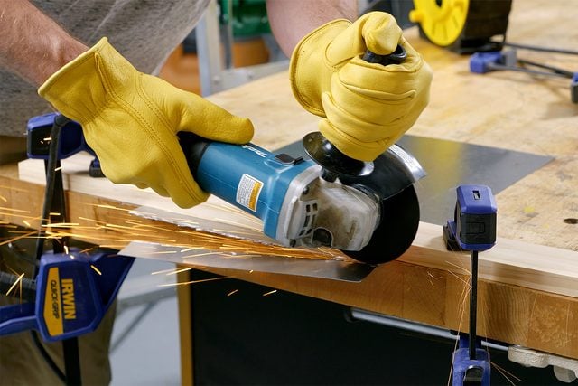 Une personne portant des gants jaunes utilise une meuleuse d’angle bleue pour couper un morceau de métal fixé à un établi. Des étincelles jaillissent du métal pendant qu'il est coupé. L'établi est équipé de pinces bleues pour maintenir le matériau en place.