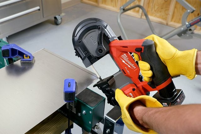 Une personne portant des gants jaunes utilise un outil électrique portatif rouge de marque Milwaukee pour couper une tôle fixée à un établi dans un atelier. L'arrière-plan comprend divers outils et machines.