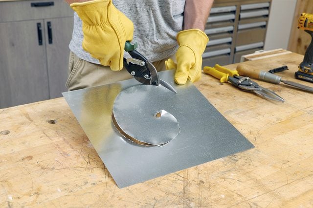 Une personne portant des gants jaunes utilise des cisailles à ferblantier pour découper une forme circulaire dans une feuille de métal sur un établi en bois. Divers outils sont éparpillés sur le banc, notamment une autre paire de cisailles, une perceuse et un ruban à mesurer.