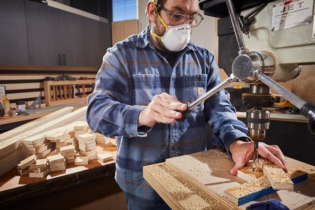 Une personne portant des lunettes de sécurité, un masque et une protection auditive utilise une perceuse à colonne pour travailler sur un morceau de bois dans un atelier, avec des morceaux de bois empilés en arrière-plan.