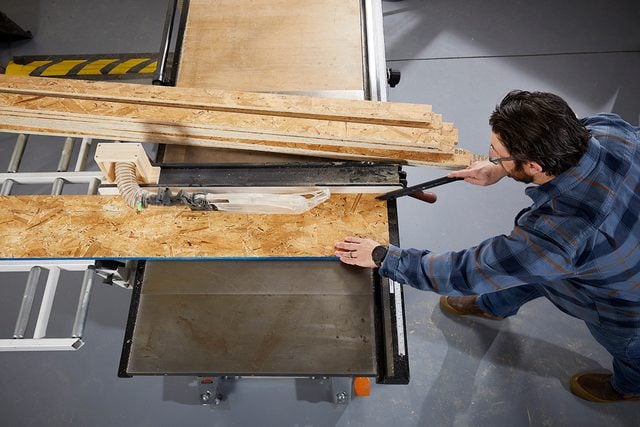 Une personne en chemise à carreaux utilise une scie à table pour couper un morceau de contreplaqué dans un atelier, avec d'autres morceaux de bois empilés à proximité.