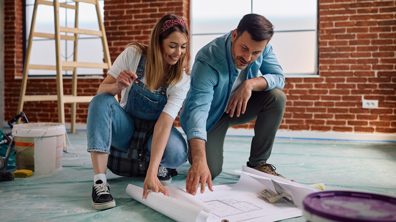 homme et femme en vêtements de travail dans une pièce en brique avec une échelle et de la peinture regardant des plans
