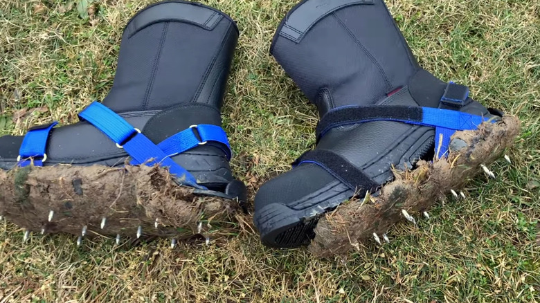 Des pointes d'aérateur à sangle sur une paire de bottes noires posées sur le sol recouvertes d'herbe accumulée.
