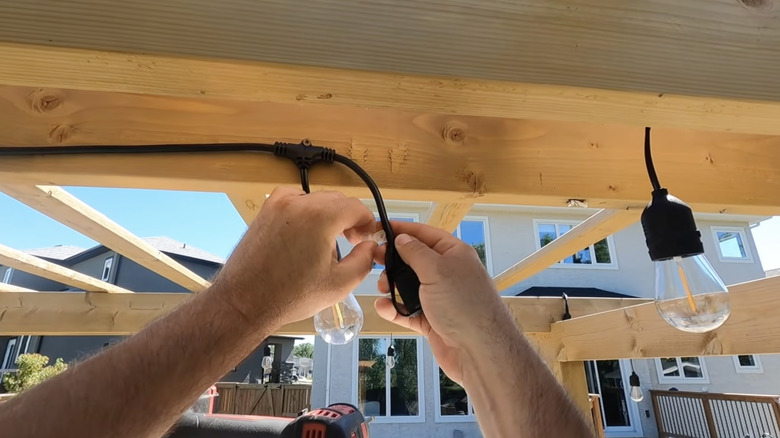 Une personne accroche des guirlandes lumineuses avec de grosses ampoules Edison sur le toit d’une pergola en bois.